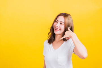 Wall Mural - Portrait Asian Thai beautiful happy young woman wear white t-shirt standing smiling white teeth, making call me gesture by finger hand isolated on yellow background, with copy space