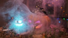 Close-up Of Two Carved Pumpkin Lanterns Rotating Amidst Thick Fog And Neon Light Standing In Courtyard Of House During The Halloween Celebration.