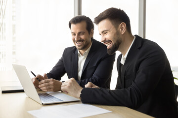 Wall Mural - Positive confident business partners men watching content on laptop together, looking at display, laughing, discussing application, software product, online startup project, smiling, laughing