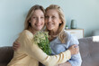 © fizkes - Happy pretty young adult daughter woman and senior blonde mom celebrating 8 march, birthday, holding bouquet of spring flowers, hugging, looking at camera, smiling. Family portrait