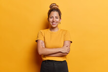 Horizontal Shot Of Lovely European Woman With Hair Bun Keeps Arms Folded Stands Satisfied Poses For Making Photo Against Vivid Yellow Background Dressed In Casual T Shirt And Black Trousers.