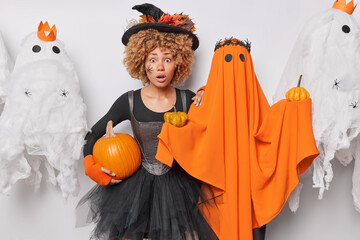 Wall Mural - Worried shocked curly haired young woman poses with pumpkin near orange ghost wearing black dress and witch hat prepares for Halloween party surrounded by spooky ghosts feels very frightened