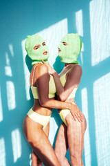 Two beautiful sexy women in green underwear. Models wearing bandit balaclava mask. Hot seductive female in nice lingerie posing near blue wall in studio. Crime and violence. Shadow from window. Hug