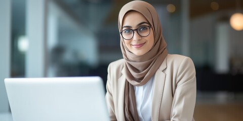 Portrait of a cheerful Muslim woman in eyeglasses wearing hijab working with laptop computer in her modern business office.