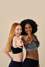 Two Happy Diverse Fit Women, African And European Young Sporty Gen Z Girls Friends Models Wearing Sportswear Pointing At You Standing Looking At Camera On Beige Background. Vertical.
