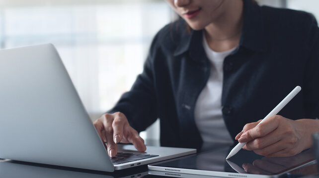 Young asian business woman using digita tbalet and working on laptop computer at modern office. Woman graphic designer, illustrator using grahic tablet, online working at office, freelance at work