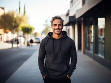 Attractive Sporty Man Dressed In A Blank Black Hoodie With Hood And Kangaroo Pocket Against The Background Of The City Street. Mockup Template For Branding Or Printing