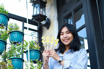 happy asian young woman hipster looking to camera with holding 2024 number candle celebrating new year eve, beautiful female standing in front of vintage house outdoor garden with low angle view