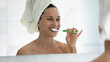 © fizkes - Smiling young woman in towel after shower look in bath mirror brush white healthy teeth with toothbrush and toothpaste, happy girl do daily facial skincare routine in bathroom, oral hygiene concept