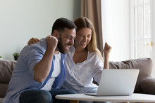 Excited Overjoyed Couple Winning Money Prize, Getting Happy Notice, Making Winner Yes Hands, Laughing, Screaming For Joy, Sitting On Sofa, Celebrating Success At Laptop At Home