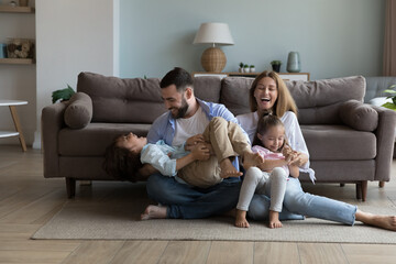 Wall Mural - Excited joyful parents tickling energetic little kids on warm floor, laughing, having fun, enjoying family activity at home. Happy mom and dad playing with giggling, shouting son and daughter kids