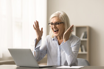 Wall Mural - Cheerful blonde elderly business woman celebrating professional success, working at laptop, laughing, shouting for joy at home office workplace, getting good news from video call