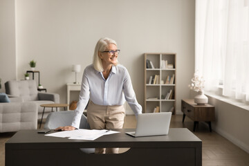 Happy successful senior freelance business woman standing at home workplace table with laptop, looking away with toothy smile, enjoying success, planning, thinking on project strategy, smiling