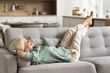 © fizkes - Joyful carefree retired woman enjoying relaxation on soft comfortable sofa, placing legs up on back, lying with closed eyes, smiling, spending weekend, leisure time at cozy home