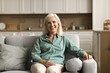 © fizkes - Positive blonde elderly woman relaxing at home, sitting on comfortable soft couch, looking at camera with toothy smile, laughing, enjoying retirement, comfort in modern apartment