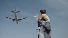 Father With A Toy Plane And A Daughter, Sitting On His Shoulders, Watching In Admiration How A Real Airplane Flying Low Overhead In A Clear Blue Sky. Concept Of A Dream About Traveling Far Away