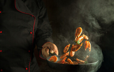 Wall Mural - Professional chef cooks shrimp in a hot pan with steam. The concept of cooking seafood or healthy vegetarian food and eating on a dark background. Freeze in motion. Free space for recipe or menu