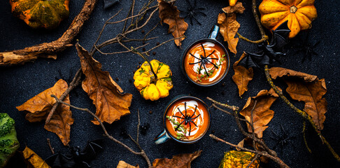 Wall Mural - Halloween pumpkin soup with cream and .thyme. Autumn festive vegan vegetarian food. Soup mug on black background with fallen oak leaves, pumpkins, spiders, twisted branches and bats. Top view