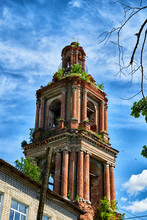 A Tall Old Stone Bell Tower Made Of Red Brick Against A Blue Sky With White Clouds And Green Trees Around. Partially Destroyed Ancient Bell Tower And Tower On A Summer Day