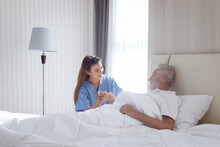 Smiling Beautiful Caring Female Doctor Holding Hand Of Male Senior Patient Who Lying In Hospital Bed. Nurse Takes Care Elderly Man Feeling Sick Need To Rest In Bed At Home, Medical Elderly Health Care