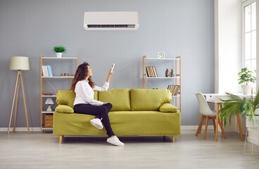 Young happy smiling woman turning on air conditioner sitting on sofa at home. Curly redhead girl homeowner enjoying cool conditioned air using remote resting on couch in the living room.