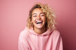 © FoxTok - Young beautiful blonde girl with curly hair in a pink hoodie laughs fervently on a pink background. Human emotions, joy, laughter, happiness. Modern student