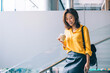 © BullRun - Happy Asian woman walking downstairs with hot drink and mobile phone