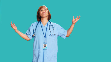 Wall Mural - Confused asian nurse frustrated about not knowing answer to patient inquiry while at work. Stumped healthcare specialist doing perplexed gesturing, isolated over blue studio background