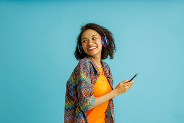 Cheerful woman in earphones with phone listen music on blue studio background and looking at side