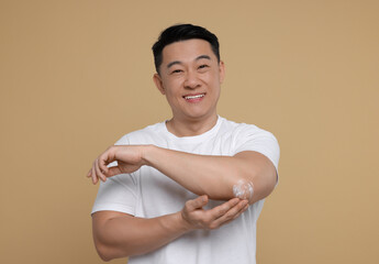 Poster - Handsome man applying body cream onto his elbow on light brown background