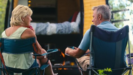 Wall Mural - Rear view of senior couple camping in countryside with RV drinking coffee by outdoor fire - shot in slow motion