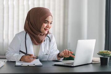 Wall Mural - Young muslim female doctor in brown hijab headscarf working with medical papers while sitting in modern clinic office.