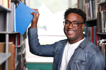 Close up smiling face young smart professor African nationality in university library, choosing book. Dark-skinned handsome guy smiling camera, taking book in blue cover from the shelf with his hand