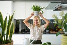 Happy Blond Woman Standing With Plant Over Head At Home