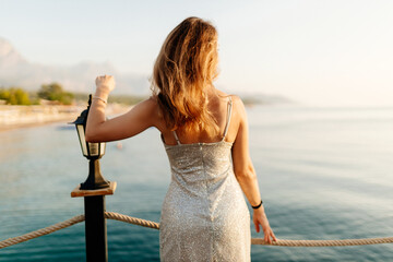 Wall Mural - A slender girl with long hair in a gray shiny dress walks along the pier by the sea at sunset. A beautiful woman in an evening dress poses against the backdrop of the ocean at dawn. Summer background