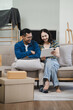 © NINENII - Young couple relaxing sitting on the sofa using the computer laptop around cardboard boxes, very happy moving to a new house.
