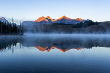 Redfish Lake In Idaho In Dawn. Calm Water Covered By Mist. Mountains Range Is Reflecting In Lake Water