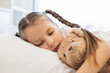 © sofiko14 - Caucasian little girl with gloomy look sleeping in white bed and seeking solace in embrace of her teddy bear. Sick kid with two braids suffering from cold and flu symptoms at home.