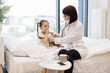 © sofiko14 - Experienced nurse sitting on bed and using stethoscope while small lady covering mouth with hand and coughing in bedroom. Concept of home treatment and friendly relations between kid and doctor.
