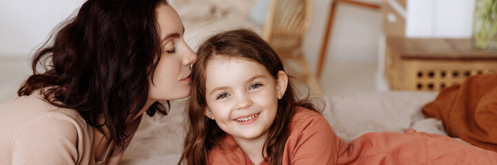 beautiful young mother kissing happy cute little girl daughter lying on bed in bedroom. mom and daug
