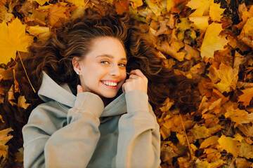 Wall Mural - Smiling young woman lying on the autumn foliage in the forest with the yellow leaves