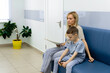 © Irina Mikhailichenko - Happy toddler boy with a woman mother in the clinic waiting for a doctor s appointment. Mom with a baby boy in a modern hospital before receiving a pediatrician. Kid 5-6 year old
