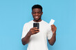 african american guy in white t-shirt uses smartphone and holds credit card on blue isolated background
