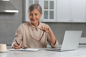 Wall Mural - Beautiful senior woman taking notes near laptop at white marble table in kitchen