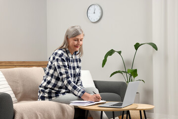 Wall Mural - Beautiful senior woman writing something in notebook while using laptop at home