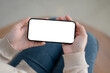 © bongkarn - A woman sitting indoors and holding a white-screen smartphone mockup in a horizontal position.