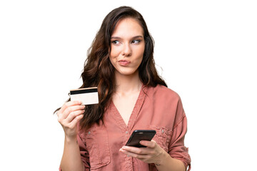 Wall Mural - Young caucasian woman over isolated background buying with the mobile with a credit card while thinking