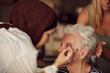 In this heartwarming real-life scene, a girl in a hijab and her sister lovingly apply makeup to their elderly grandmother, preparing her for a special family anniversary celebration, showcasing the