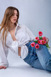 Studio portrait of a beautiful sexy slim young caucasian brown-haired girl in white shirt and blue jeans with silver chain necklace sitting and holding a bouquet of pink tulips