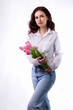 Studio portrait of a beautiful sexy slim young caucasian brunette in white shirt and blue jeans standing with a bouquet of pink tulips